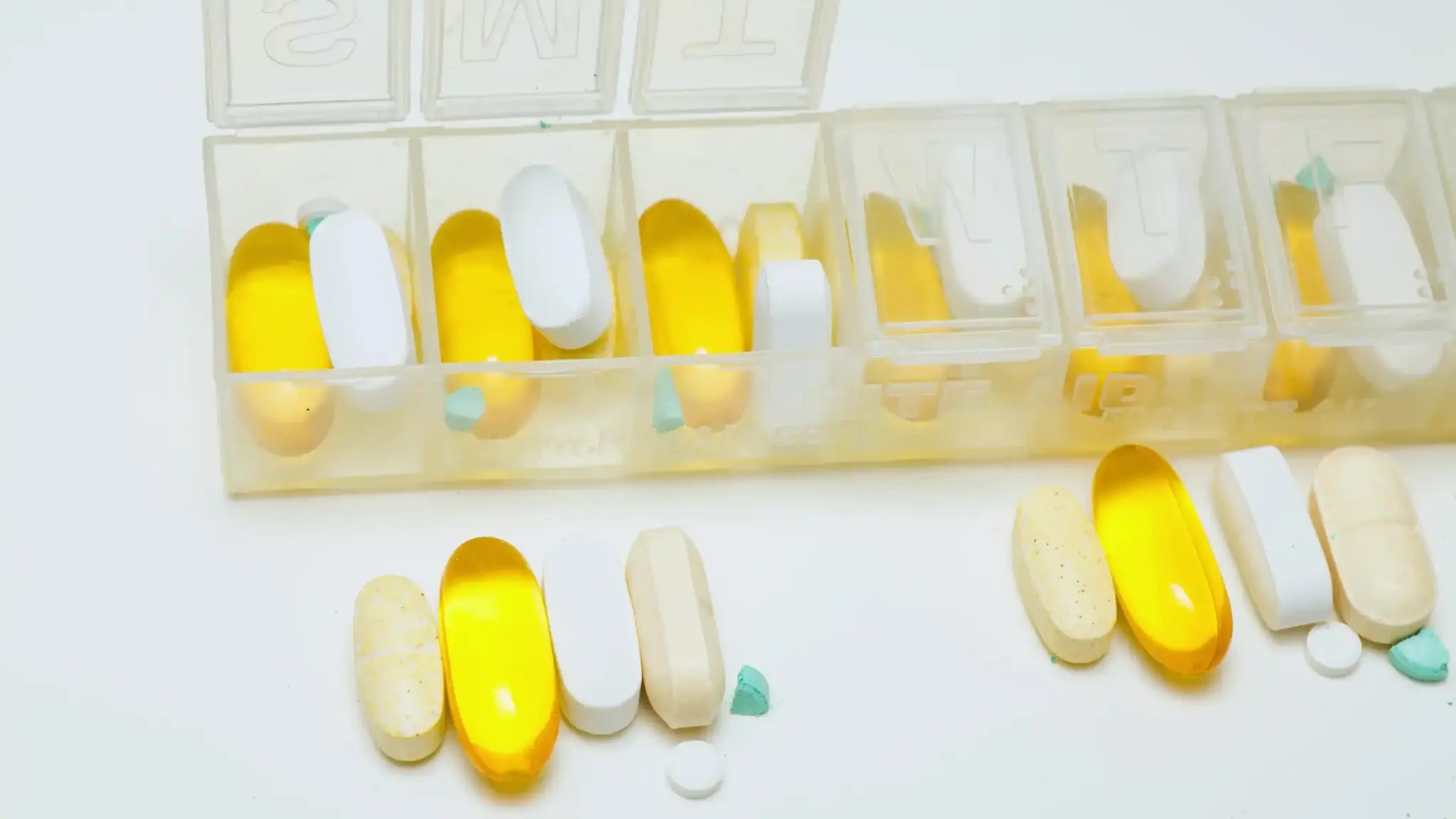 Weekly pill organizer drawer open showing yellow softgels, white tablets, beige capsules, and green fragments in individual compartments, with matching pills lined up in front, illustrating an NMN dosage schedule.