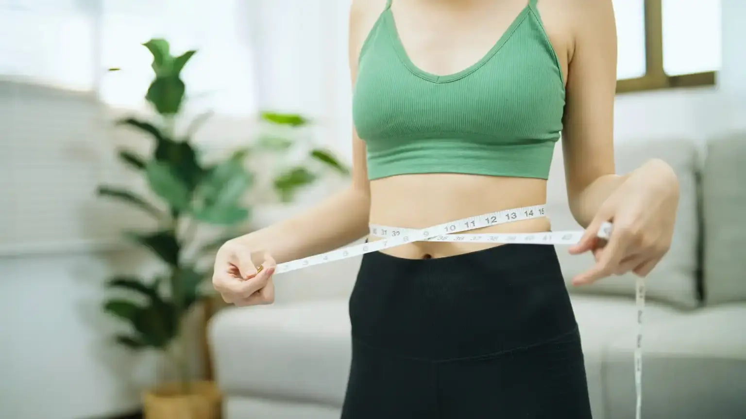 Woman in athletic wear measuring her waist with a tape measure in a bright room, illustrating berberine’s role in weight loss and blood sugar management.