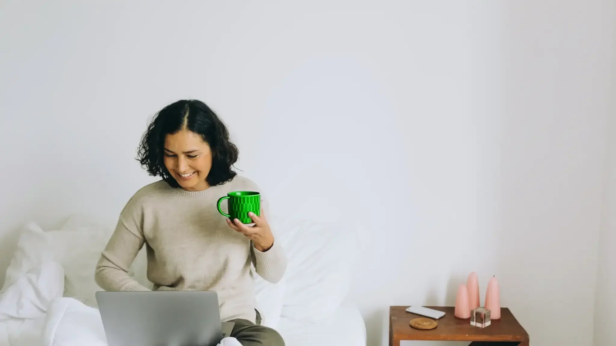 A smiling woman sits cross-legged on a cozy bed, holding a green mug in one hand as she works on a laptop, illustrating the intersection of daily habits and genetic factors in longevity and DNA-driven lifespan insights.