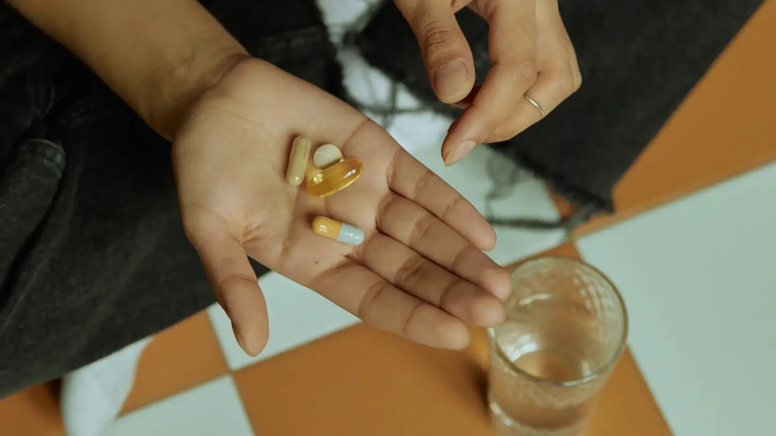 Close-up of a hand holding assorted supplement capsules—including a golden NMN softgel and bi-colored pill—over an open palm with a glass of water nearby on a tiled floor, representing Andrew Huberman's NMN insights.