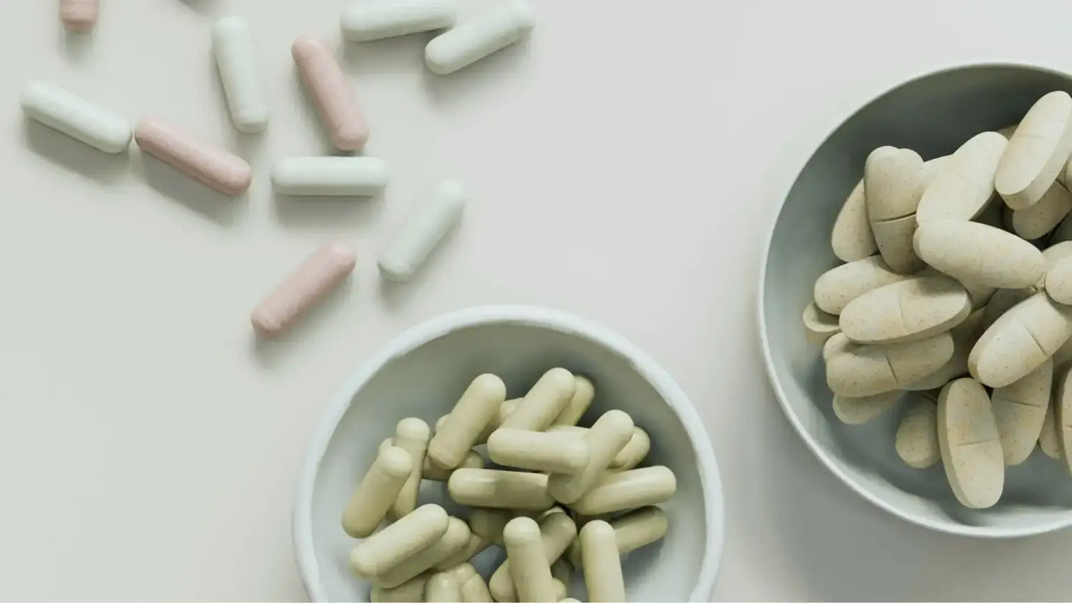 Top-down view of two bowls—one filled with beige NMN capsules, the other with oval beige tablets—and scattered pink and white capsules on a white surface, illustrating NMN supplementation timelines.