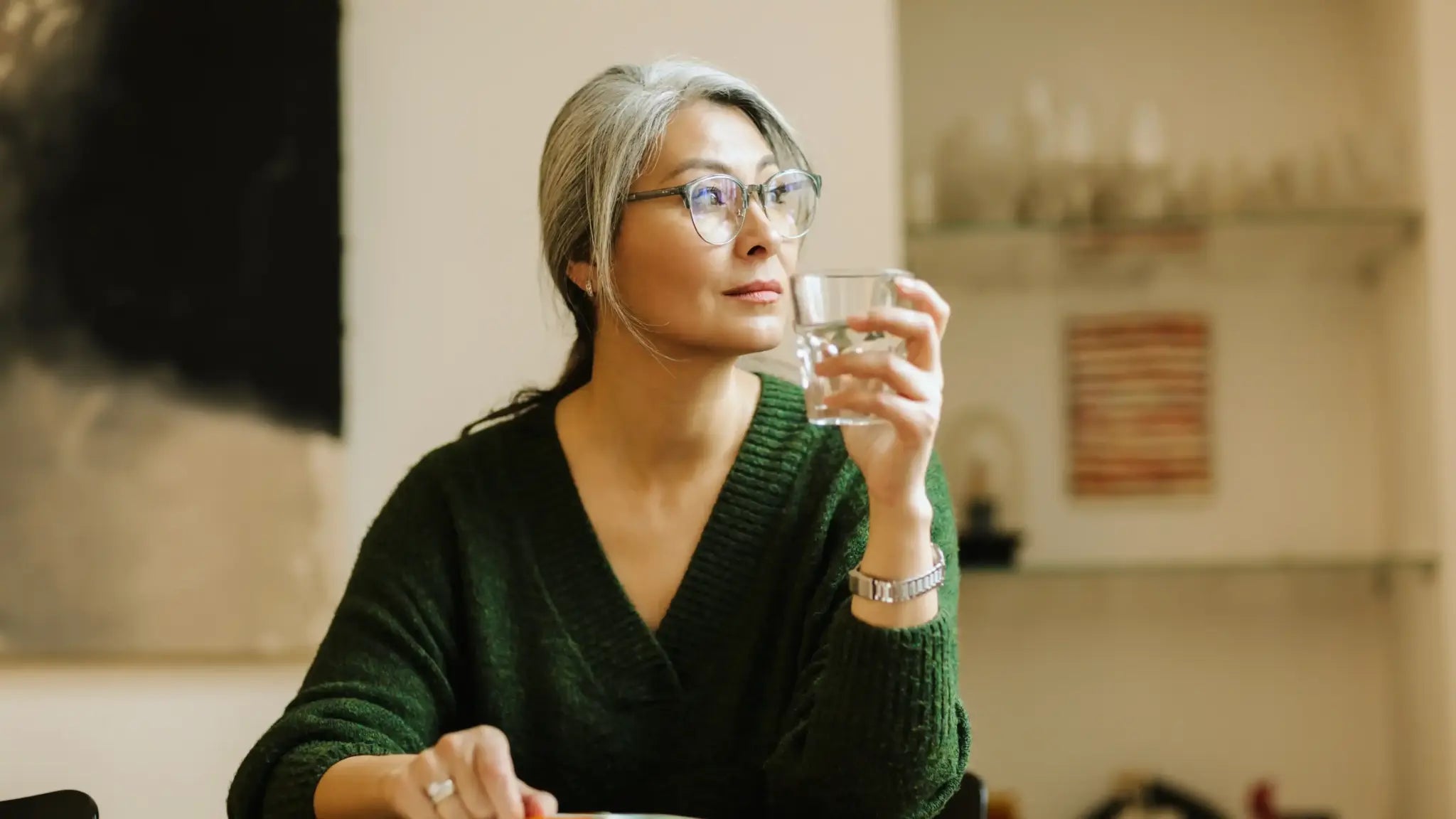Woman sitting at a table holding a glass of water and looking thoughtfully ahead, illustrating unlocking the health benefits of calcium alpha-ketoglutarate.