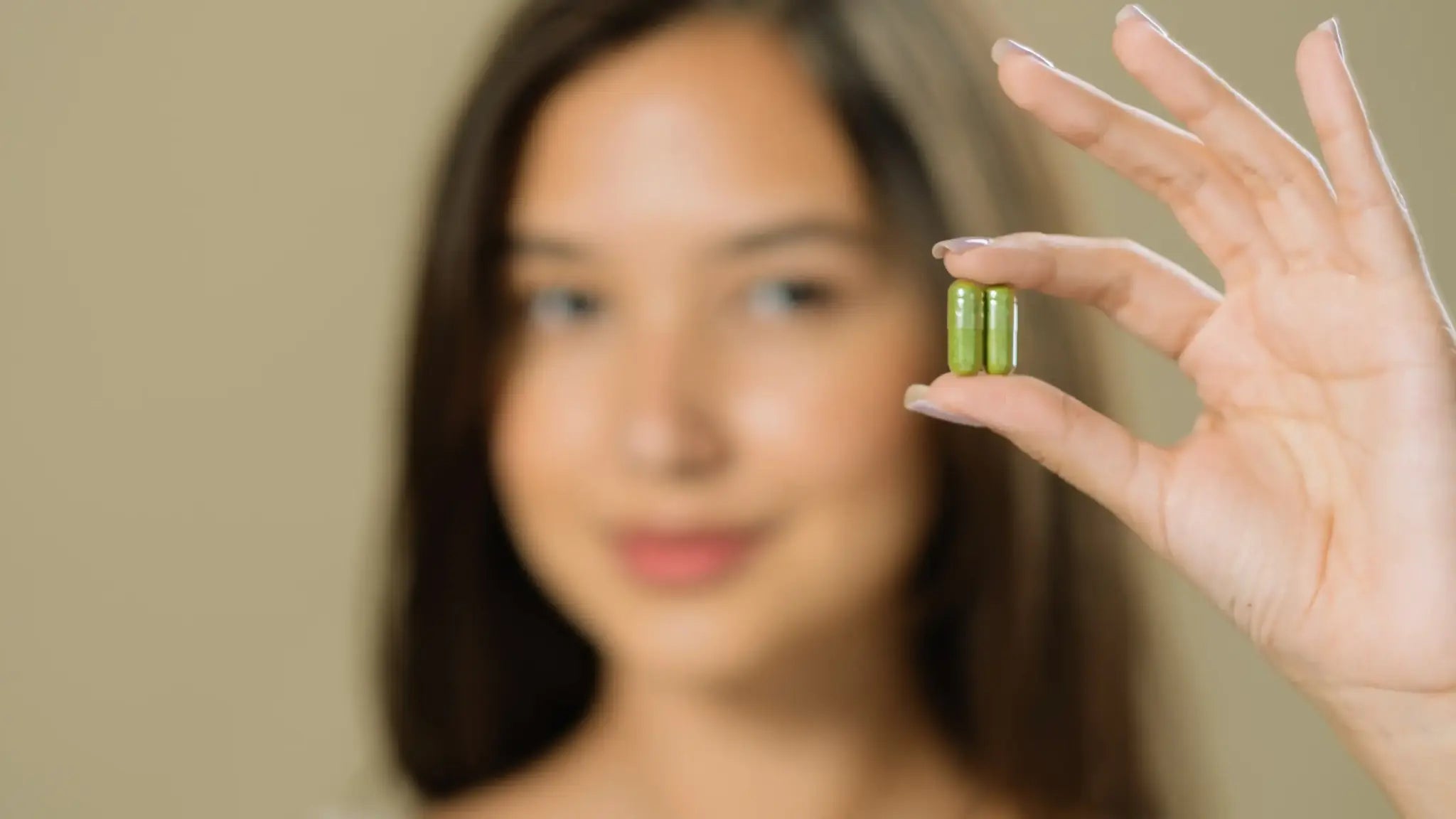 Blurred portrait of a woman holding two green capsules between her thumb and forefinger against a neutral background, symbolizing a quick comparison between partiQlar and Elysium supplements.