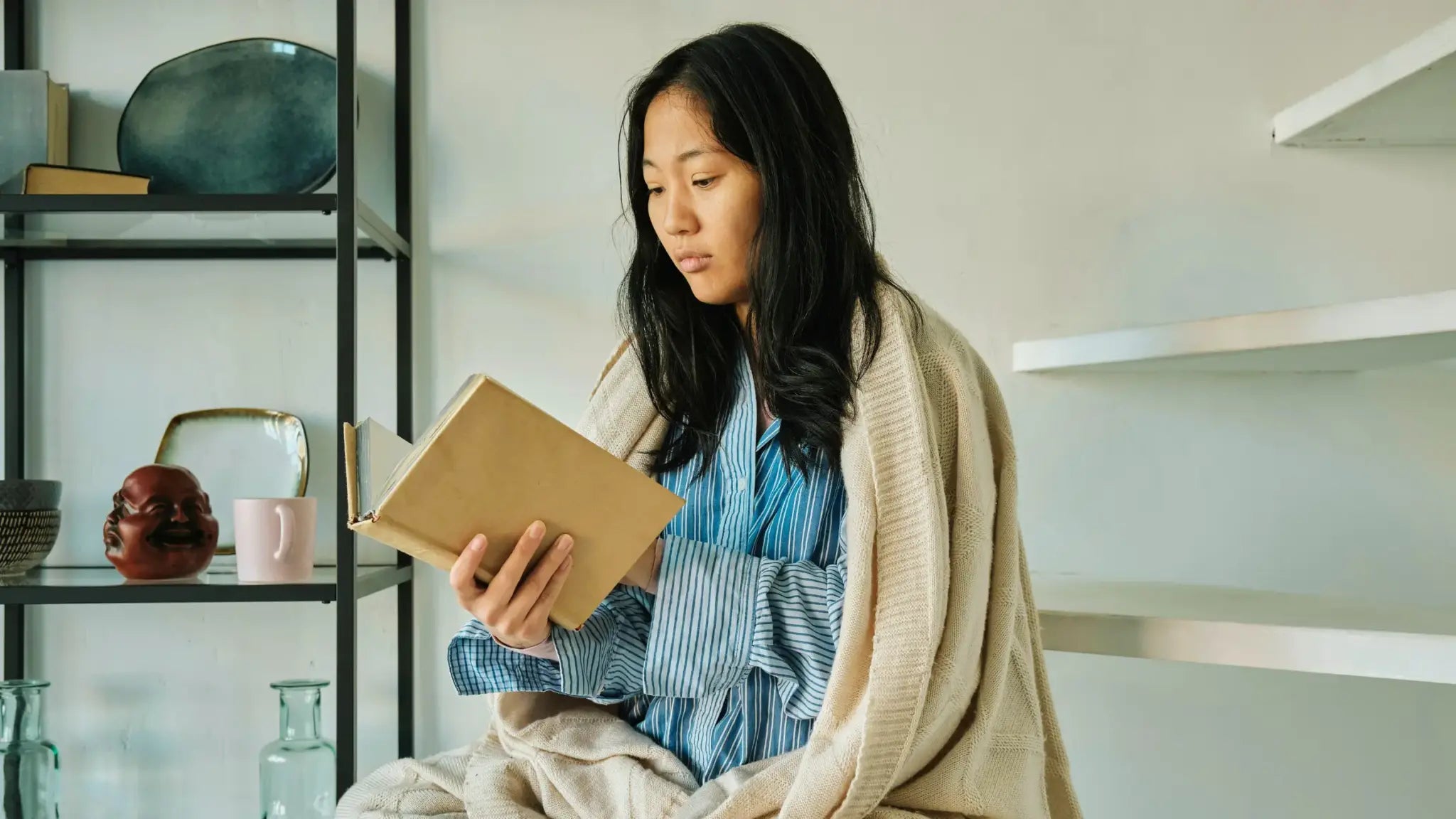 Young woman wrapped in a cozy blanket reading a book by a bookshelf, representing focus and calm as a natural Adderall alternative.