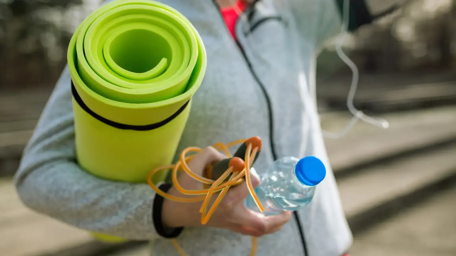 Close-up of a person carrying a rolled green yoga mat, orange jump rope, and water bottle outdoors, symbolizing fitness and hydration for TMG dosage recommendations.