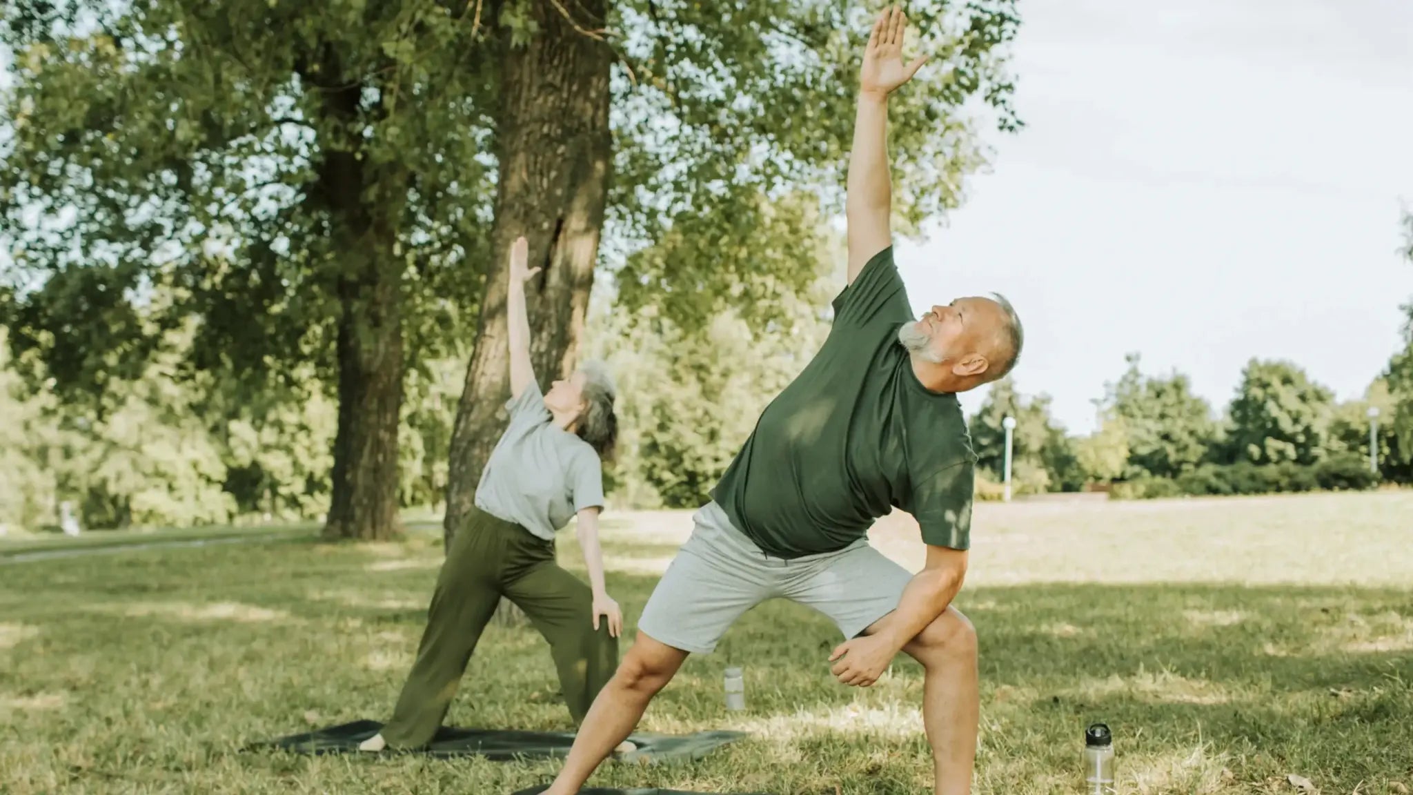Two older adults—one man and one woman—stretching their arms overhead and bending to the side while practicing yoga-like poses on mats in a sunlit park.