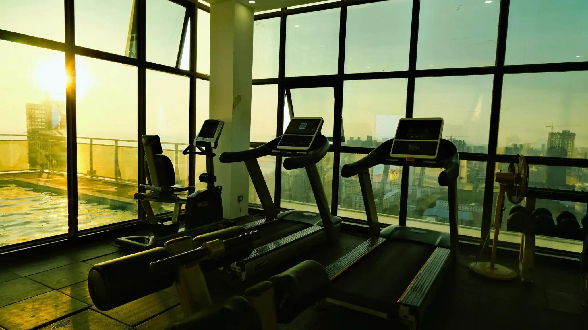 Empty high-tech gym with treadmills and exercise bikes facing floor-to-ceiling windows overlooking a city skyline and sunrise pool, illustrating Bryan Johnson’s longevity blueprint.