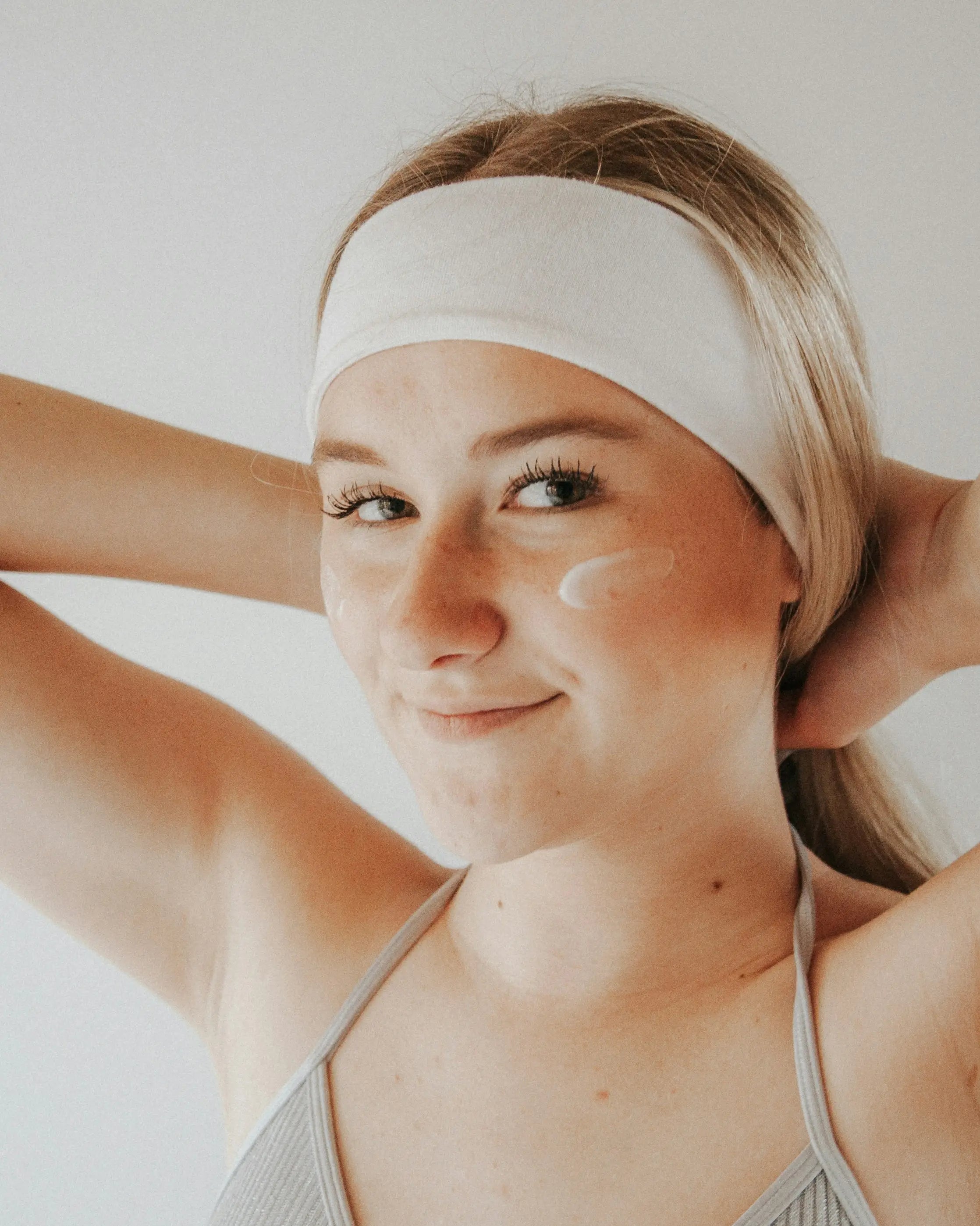 A smiling young woman wearing a headband gently applies a swipe of cream beneath her eye, illustrating glutathione’s role in brightening skin, neutralizing free radicals, and supporting overall detoxification and longevity benefits.