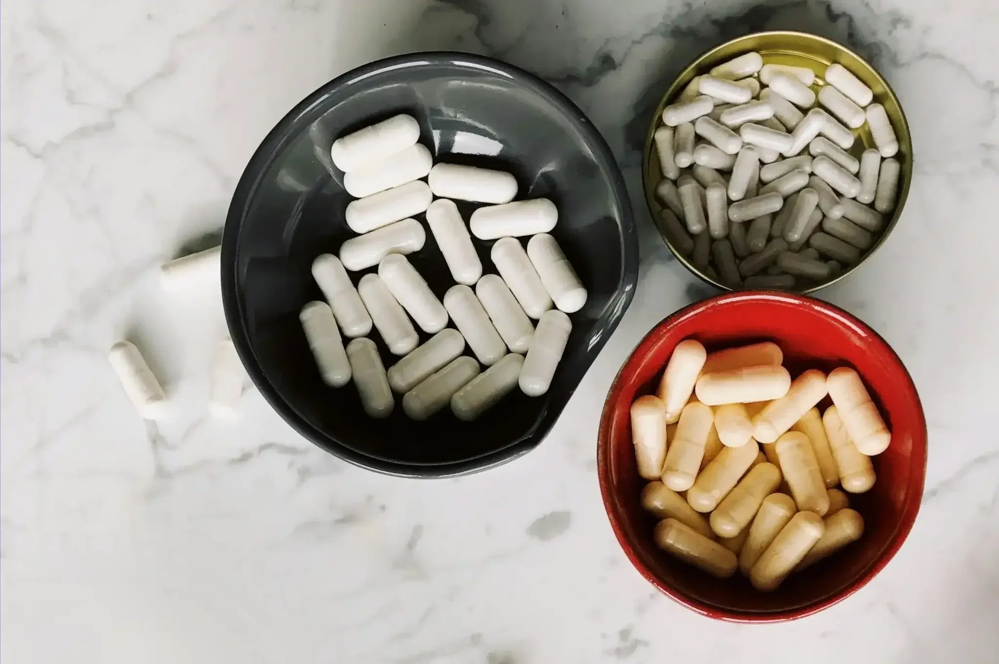 Top-down view of three bowls—gray, gold, and red—each filled with different colored supplement capsules on a marble countertop, illustrating TMG supplements with various benefits, dosages, and potential side effects.